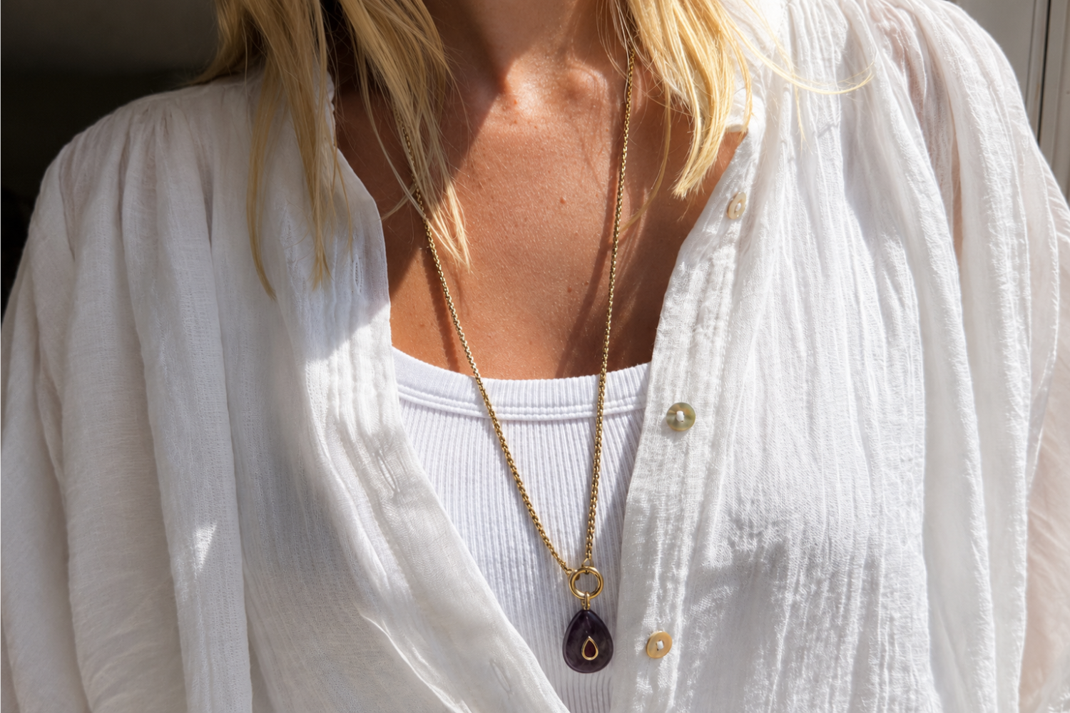 A close-up of a woman wearing a long gold chain necklace with a dark teardrop pendant over a white ribbed tank top and a loose white button-up shirt.