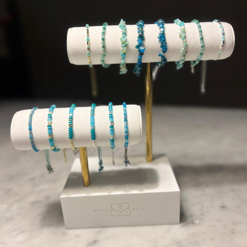 Boho Betty two-tier display stand showcasing Aqua and Turquoise gemstone bracelets on white faux suede bars with gold-tone stands, set on a marble surface.